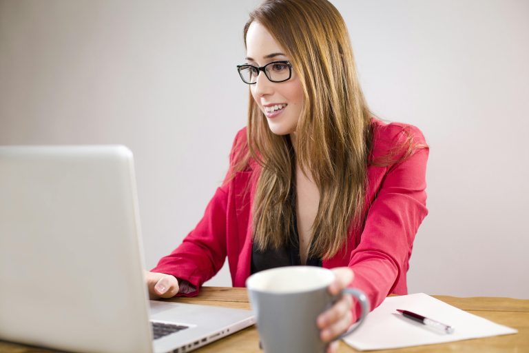 A smiling young businesswoman in a red jacket works on her laptop while holding a coffee cup.